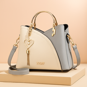 Two-tone cream and light gray leather handbag with curved panel stitching, gold hardware, heart-and-pearl tassel charm, and detachable strap, styled on a beige platform with a soft pastel gradient background.