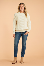 Off-white cable-knit sweater with intricate braided and diamond patterns, paired with medium-wash skinny jeans and beige suede ankle boots.