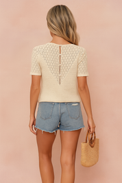 Back view of a stylish summer blouse featuring a cream crochet lace V-panel with delicate fabric buttons, paired with light-wash denim shorts and a woven straw handbag, set against a pastel sand and pink backdrop in soft studio lighting.