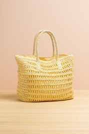 Cream-colored crochet tote bag with open-weave design and braided handles, set on a light wood platform against a softly lit beige and pastel pink gradient background.