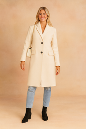 Elegant cream long coat with two-button front closure, tailored fit, and flap pockets, styled with light blue jeans and black ankle boots.