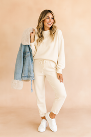 Cream lounge co-ord set featuring a relaxed-fit sweatshirt and drawstring joggers, styled with a fur-trimmed denim jacket and white sneakers for a cozy, casual look.
