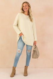 Oversized cream long-sleeve tunic paired with light blue distressed skinny jeans, taupe ankle boots, and a matching taupe handbag.