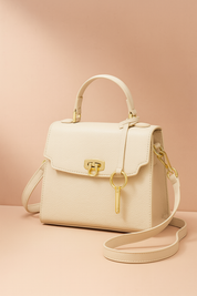 Elegant cream-colored structured handbag with pebbled texture, gold turn-lock clasp, matching top handle and adjustable strap, and a slim gold charm detail, styled on a warm blush-pink background with soft gradient lighting.