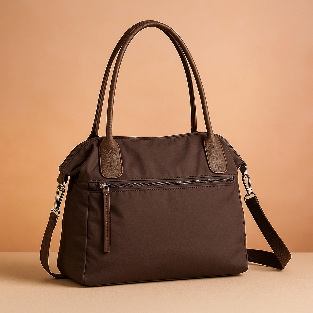 Dark brown nylon shoulder bag with smooth matte texture, two long handles, front zip pocket, and detachable shoulder strap, displayed against a soft warm sand and pastel pink gradient background.