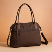 Dark brown nylon shoulder bag with smooth matte texture, two long handles, front zip pocket, and detachable shoulder strap, displayed against a soft warm sand and pastel pink gradient background.