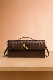 Dark brown woven leather handbag with gold knot clasp, top handle, and wrap-around strap, displayed on a neutral table with a softly lit warm blush background.