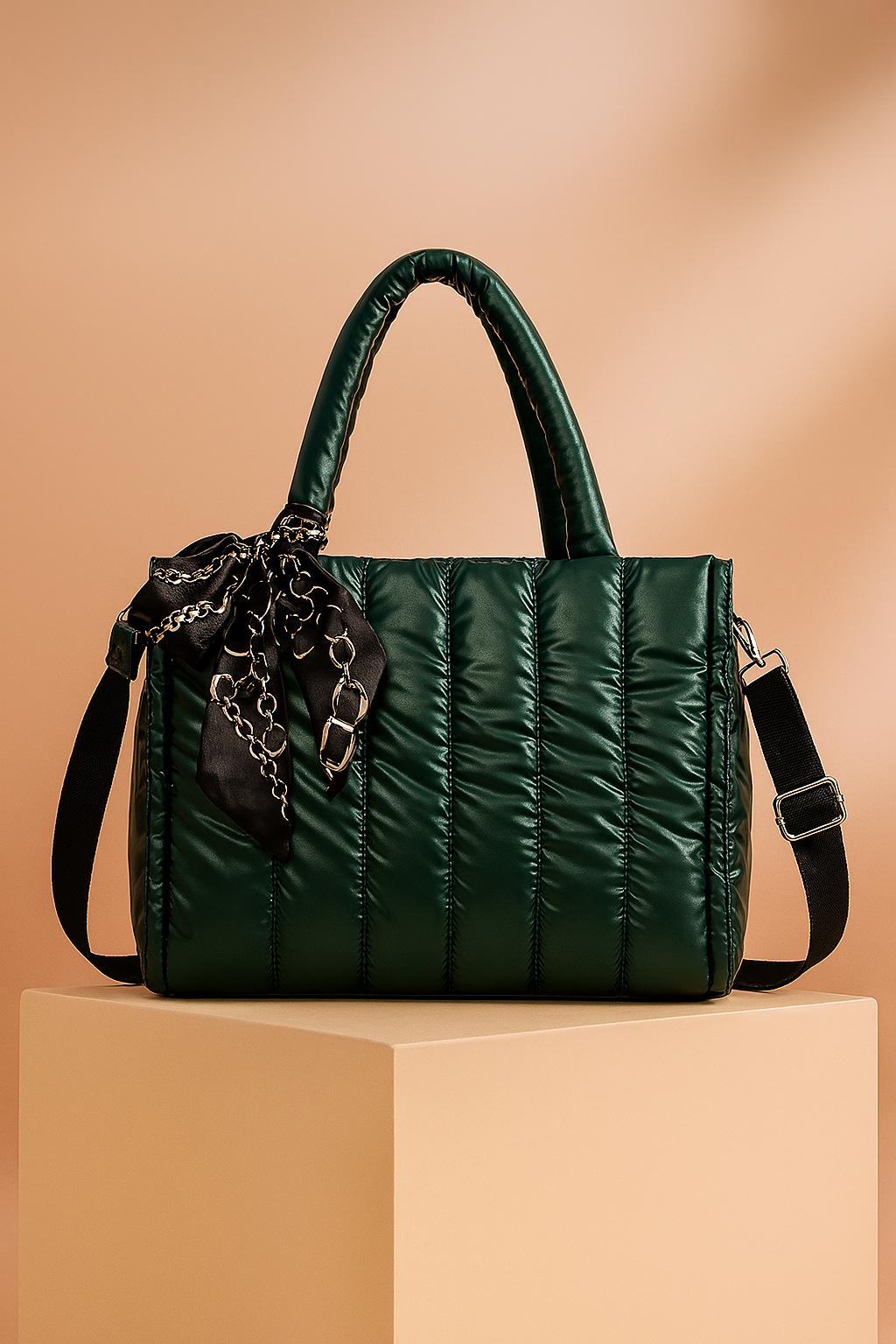 Dark green quilted satin tote bag with top handles, black adjustable strap, and black chain-print scarf tied at the handle, displayed on a pedestal with warm gradient lighting.