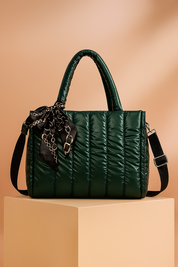 Dark green quilted satin tote bag with top handles, black adjustable strap, and black chain-print scarf tied at the handle, displayed on a pedestal with warm gradient lighting.