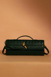 Dark green rectangular woven handbag with a top handle, gold key-shaped clasp, and crossbody strap, styled against a warm gradient backdrop.