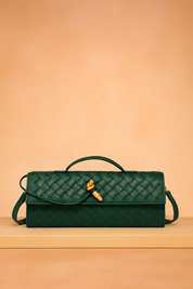 Dark green woven leather handbag with gold hardware and top handle, displayed on a light wood platform against a soft beige and pastel pink gradient backdrop.