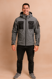 Dark grey tactical softshell jacket with black chest pockets, multiple zippers, and adjustable hood, styled with black jeans and grey sneakers.