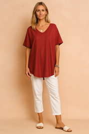 Elegant deep red short-sleeve tunic blouse made from lightly textured fabric, featuring a relaxed drape and a scoop neckline, paired with crisp white ankle-length trousers and minimal slide sandals—perfect for effortless casual chic.