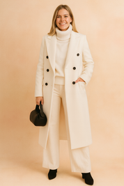 Elegant long double-breasted coat with button closure, styled over a turtleneck sweater and wide-leg trousers, paired with black ankle boots and a handbag.