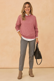 Dusty pink knit sweater layered over a white collared shirt, paired with gray tailored trousers and taupe suede ankle boots, accessorized with a black leather handbag.
