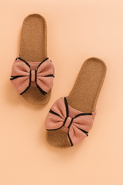 Women's flat slide sandals with large dusty rose fabric bows accented by bold black trim, featuring a woven natural-look footbed with tight-knit straw texture.