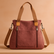 Dusty rose canvas tote bag with tan faux leather accents, dual zipper pockets, and adjustable brown shoulder strap on a warm gradient background.