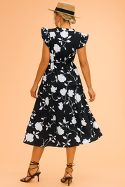 Back view of a tall woman in a black floral summer dress with flutter sleeves, wearing a straw hat and gold accessories, posed against a softly lit warm sand and pastel pink background in fashion photography style.