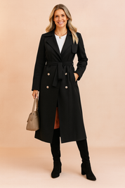 Elegant black double-breasted trench coat with matching belt, silver-tone buttons, and side pockets, styled over a white top and paired with black knee-high boots and a taupe handbag.