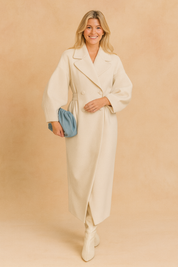 Long cream double-breasted coat with wide lapels, puff sleeves, and belted waist paired with white boots and a blue clutch.