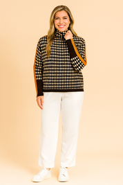 Oversized houndstooth turtleneck sweater with bold orange stripe detailing on sleeves, paired with black jogger pants and white sneakers.