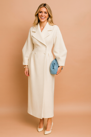 Elegant long off-white double-breasted wool coat with wide lapel collar, structured puff sleeves, and a fitted waistline, paired with a light blue ruched clutch and cream pointed-toe heels.