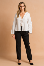White V-neck blouse with long sleeves and double front tie closure, styled with black tailored trousers and black heels.