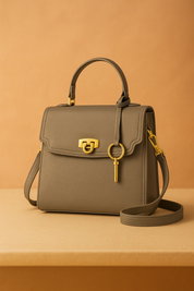 Elephant gray pebbled leather handbag with gold hardware, scalloped flap closure, key charm, and top handle, styled on a table in softly lit studio setting.