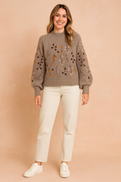 Taupe-gray knit sweater with floral embroidery in yellow, brown, and blush tones, paired with cream straight-leg trousers and white sneakers.