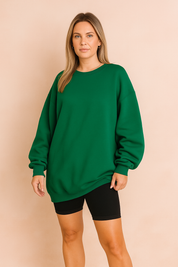 Oversized emerald green crewneck sweatshirt for women with dropped shoulders and ribbed cuffs, styled with black biker shorts in a studio with warm-toned pastel backdrop.