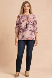 Loose-fit floral blouse with watercolor-effect purple and orange flower motifs on a pale pink base, featuring 3/4 sleeves and a straight hem, styled with dark jeans and taupe heels.