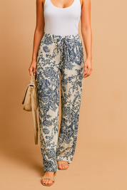Wide-leg cream pants with intricate navy blue floral print, a drawstring waist, and a relaxed fit styled with beige sandals and a tan shoulder bag.
