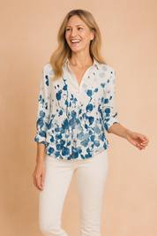 White collared blouse with blue and navy floral gradient print concentrated toward the bottom and sleeve ends, featuring a classic button-down front and 3/4 rolled sleeves, paired with white fitted trousers and a brown leather belt.