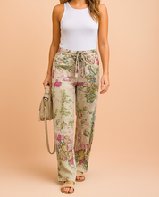 Wide-leg drawstring pants featuring a soft beige base with intricate green foliage and magenta floral prints, styled with a ribbed white tank and beige sandals for a breezy casual look.