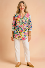 Colorful floral V-neck blouse with three-quarter sleeves, featuring a vivid multicolor garden print in pink, green, blue, and yellow tones; styled with white pants and tan block-heel shoes.