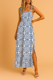 A European woman modeling a sleeveless blue and white summer dress with bow straps and a thigh-high slit, set against a soft warm tan background.
