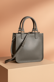 Structured gray leather handbag with two top handles, clean edge stitching, and a matching long shoulder strap with silver-tone hardware, presented against a soft warm-toned background.