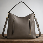 Gray leather hobo bag with gold hardware and dual straps, displayed on a wooden platform with a soft warm gradient backdrop.