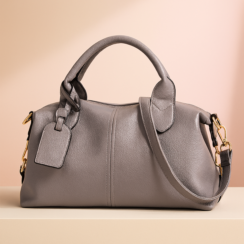 Gray pebbled leather satchel handbag with dual handles, gold-tone hardware, and side strap, styled on a soft neutral platform with a warm sand and pastel pink gradient backdrop.