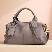 Gray pebbled leather satchel handbag with dual handles, gold-tone hardware, and side strap, styled on a soft neutral platform with a warm sand and pastel pink gradient backdrop.
