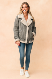 Gray shearling-lined aviator jacket with oversized lapel collar, zip closure, adjustable buckle straps on cuffs and hem, side pockets, and soft faux fur lining, styled with fitted blue jeans and white sneakers.