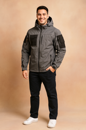 Gray tactical softshell jacket with black utility pockets, multiple zippers, and a hood, styled with black trousers and white sneakers.