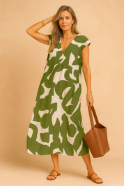 European woman modeling a green and beige abstract print summer dress with V-neckline, styled with a brown leather tote and sandals, standing in a warm sand and pastel pink studio background.