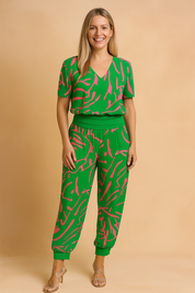 Two-piece coord set featuring a vibrant green base with bold hot pink abstract brushstroke pattern, including a short-sleeve V-neck top and relaxed-fit jogger-style pants with a wide waistband and cuffed ankles. The fabric showcases a fluid drape and consistent print saturation, maintaining the original design's exact color shades and graphic pattern placement.