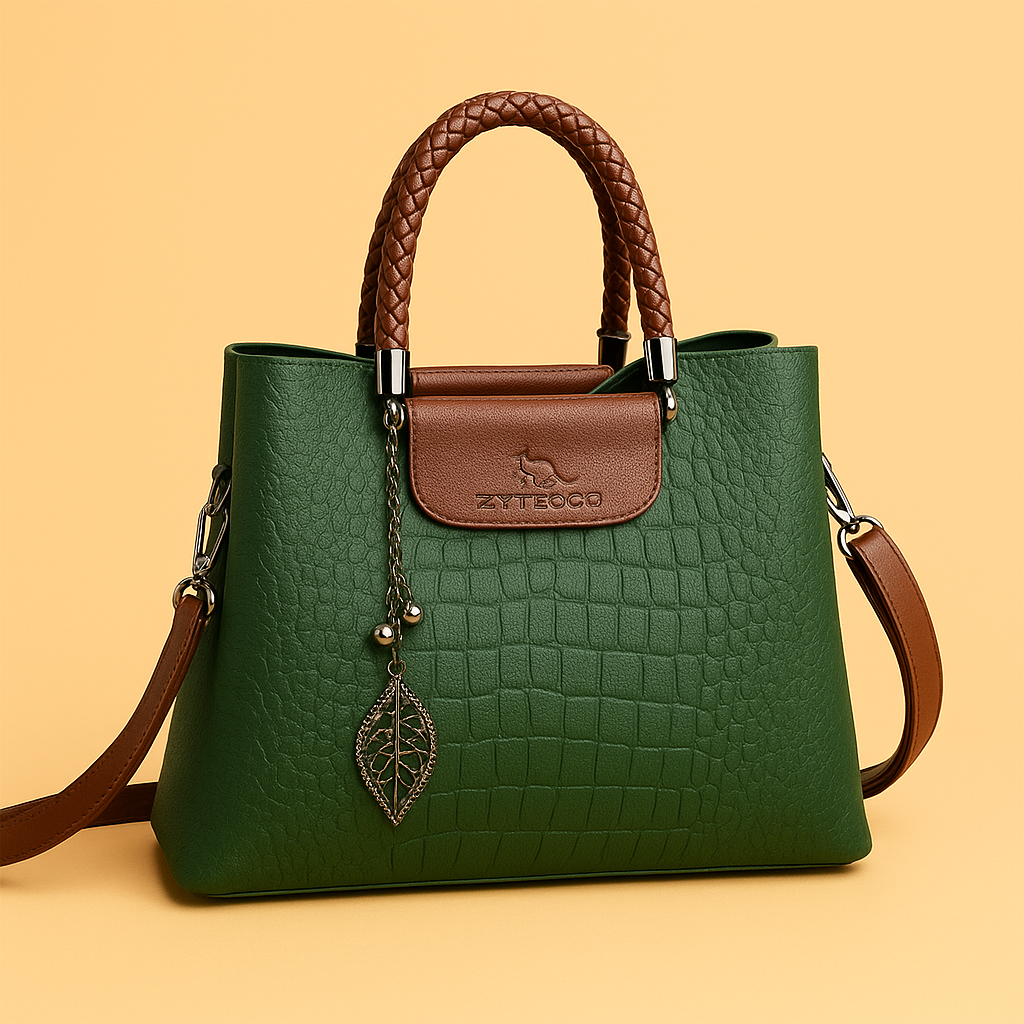 Elegant crocodile-textured green handbag with braided brown handles, brown flap with embossed logo, and metallic leaf pendant, styled cleanly against a warm sand-toned background.