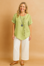 Light green short-sleeve linen blouse featuring a scoop neckline and diagonal row of brown buttons, styled with off-white wide-leg pants and a black pendant necklace.