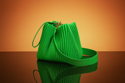 Green pleated leather handbag with gold hardware and long strap, placed on a glossy surface with warm gradient backdrop.
