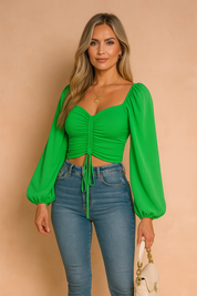 A fashion model with long ash blond hair wearing a vibrant green ruched crop top with balloon sleeves and high-waisted jeans, posing against a softly lit warm sand and pastel pink background in a studio setting.