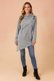 Long-sleeve heather grey asymmetrical tunic with crossover front design, high cowl neck, and three decorative black buttons, paired with slim blue jeans and black ankle boots.