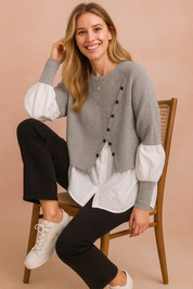 Layered outfit featuring a cropped grey buttoned knit sweater worn over a long white shirt with puff sleeves, paired with straight black trousers and white lace-up sneakers.